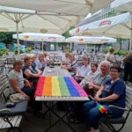 Mehrere Omas sitzen um einen langen Tisch im Freien, auf dem Tisch liegt ausgebreitet eine Regenbogenfahne